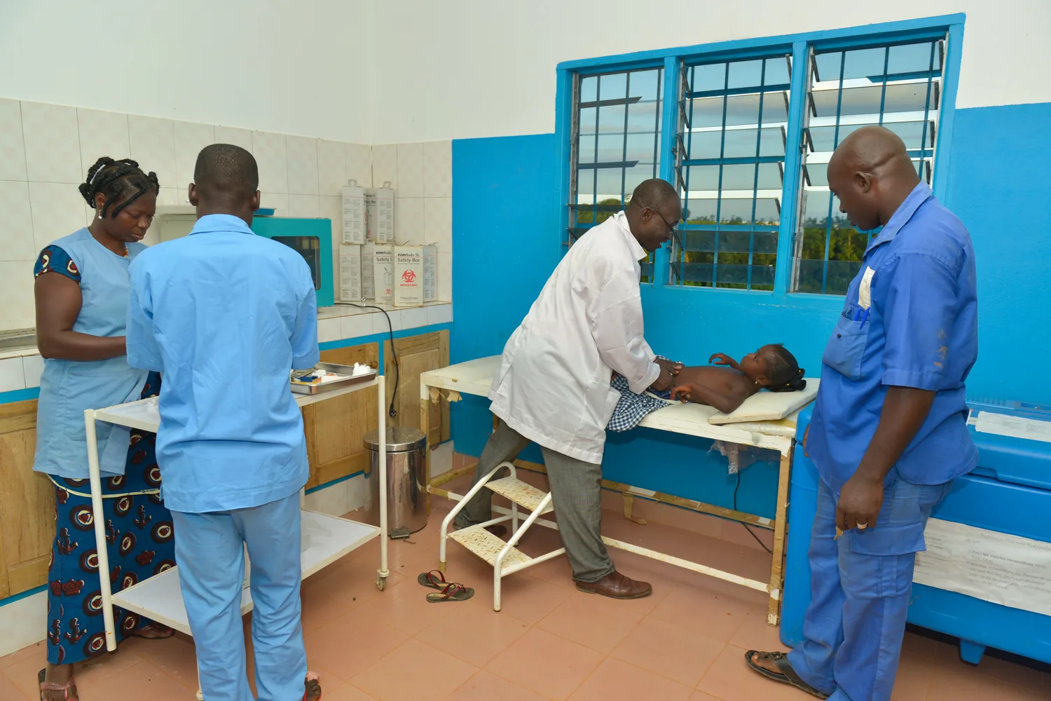 Africa photo — Un médecin en blouse blanche examine un patient allongé sur une table de consultation, entouré de trois autres professionnels de santé