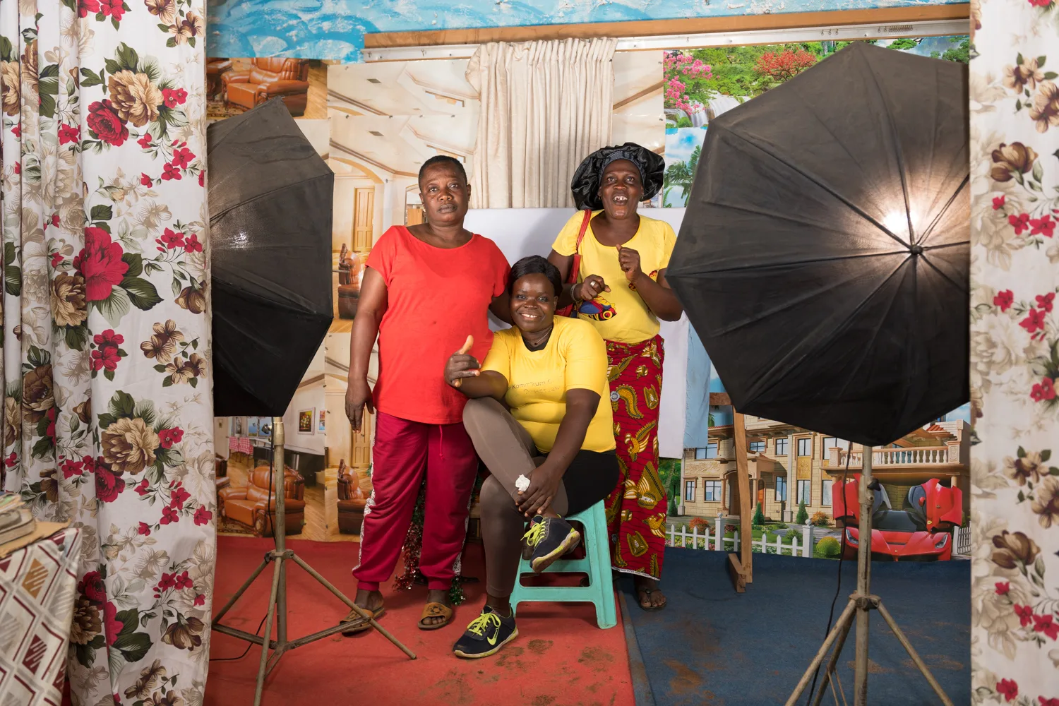Africa photo — Trois femmes souriantes posant devant un fond décoratif floral et des parapluies de studio photo, avec une ambiance chaleureuse et arti