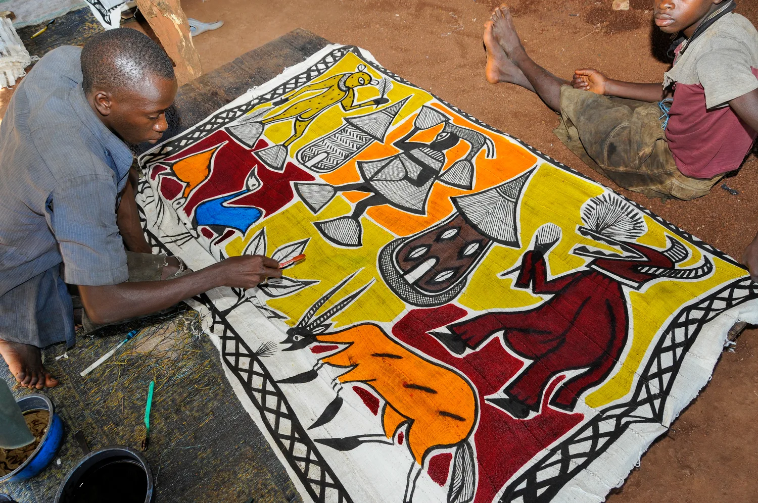 Africa photo — Un artisan s’affaire sur un grand tissu étendu au sol, richement décoré de motifs géométriques et de représentations d’animaux dans des