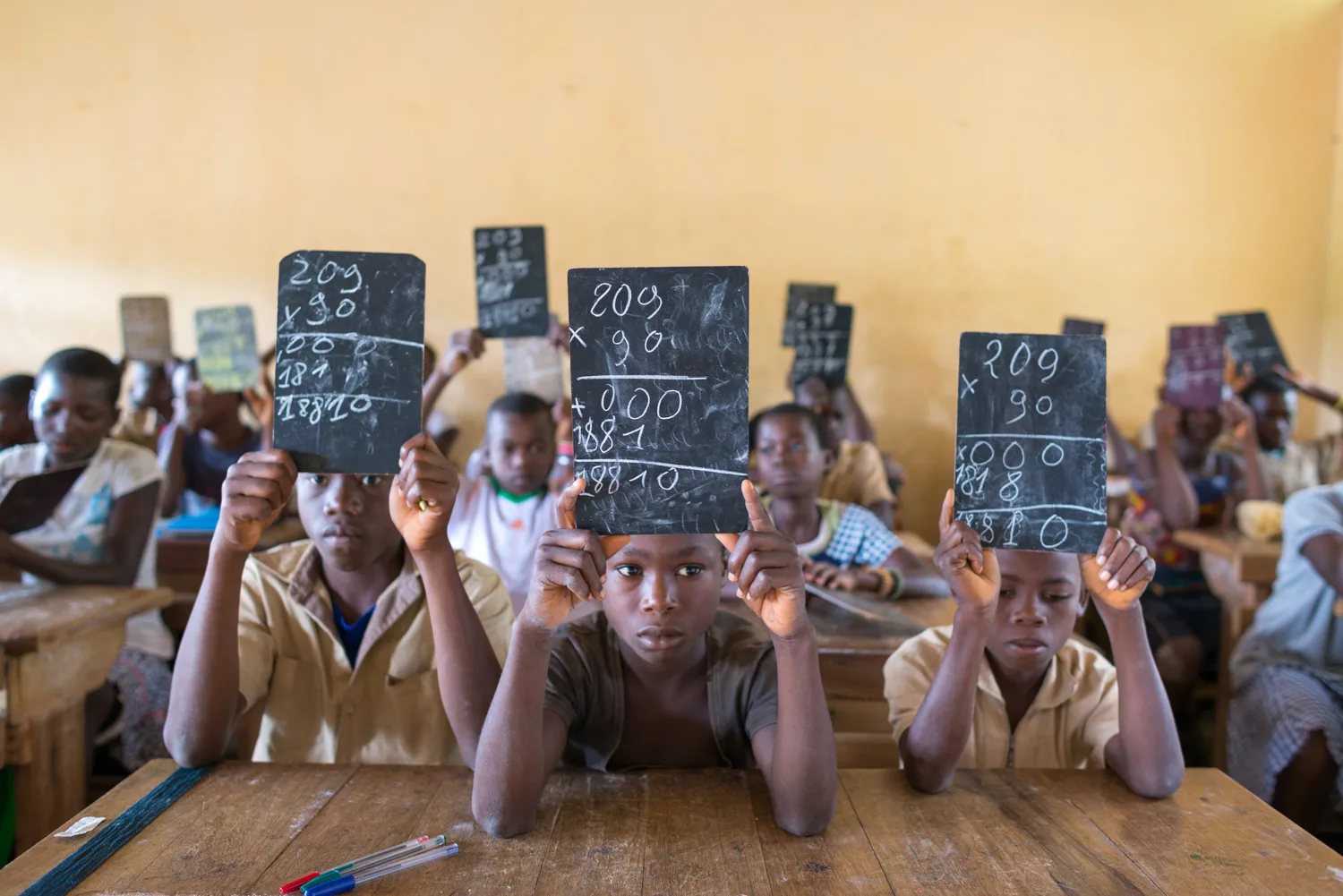 Africa photo — Trois enfants en classe lèvent des ardoises noires où sont écrits des calculs mathématiques, notamment 209 multiplié par 90 avec les ét
