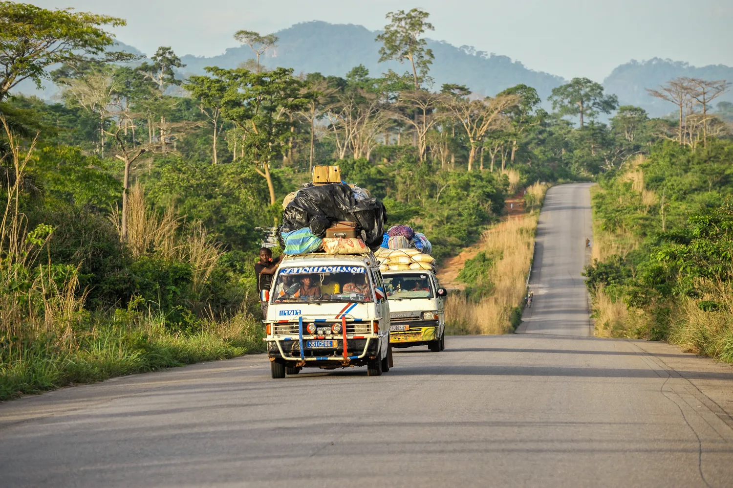 Africa photo — Deux véhicules utilitaires remplis de sacs et de marchandises circulent sur une route asphaltée bordée de végétation dense, sous un cie