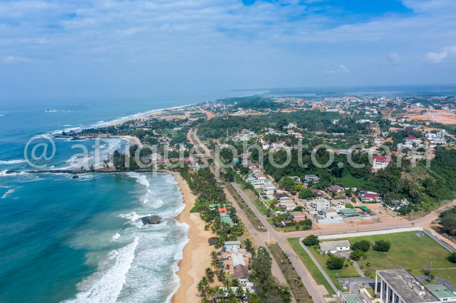 Africa photo — Une vue aérienne d’une longue plage de sable fin bordée d’une eau turquoise et de vagues douces. À droite, une route sinueuse longe des