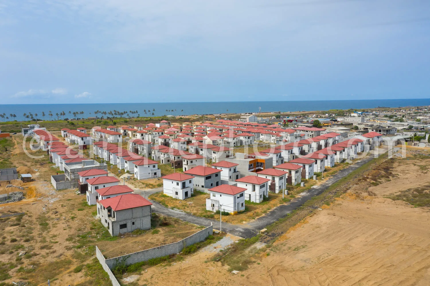 Africa photo — Vue aérienne d'un lotissement de maisons en construction avec des toits rouges, situé en bordure de plage, avec un chemin central et de