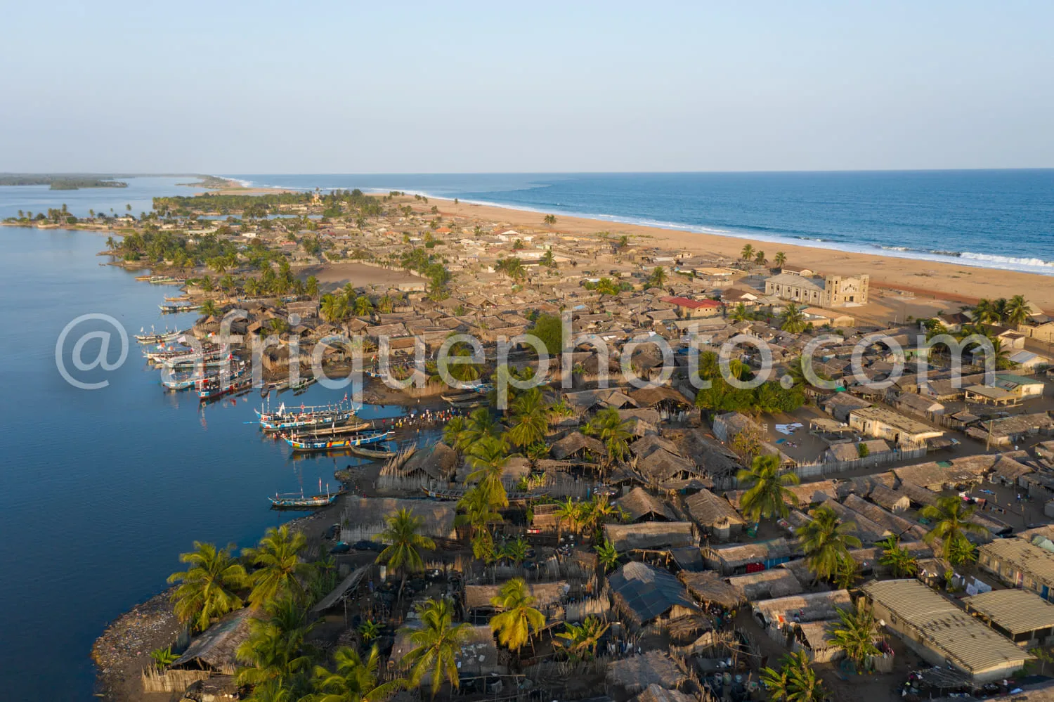 Africa photo — Vue aérienne d’un village côtier africain où l’on distingue des maisons en toiture de paille ou de palme, une lagune bordée de bateaux