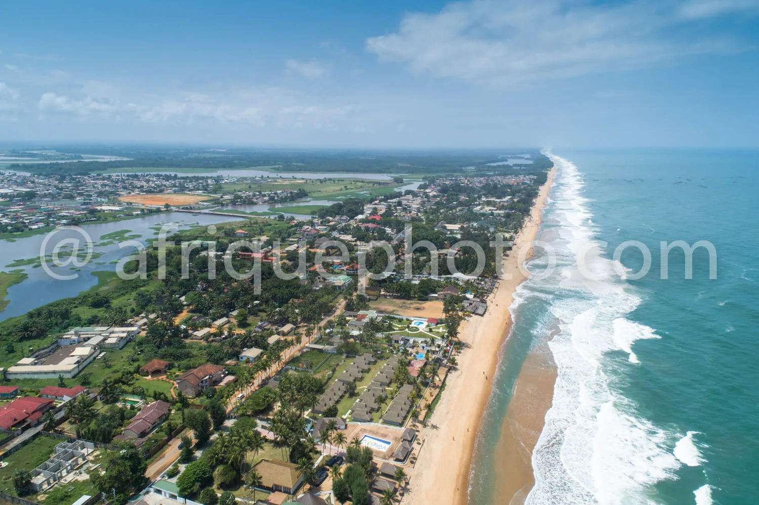 Africa photo — Image montrant une longue plage de sable fin bordée d’un océan aux vagues bleutées. À gauche, une végétation dense avec des habitations