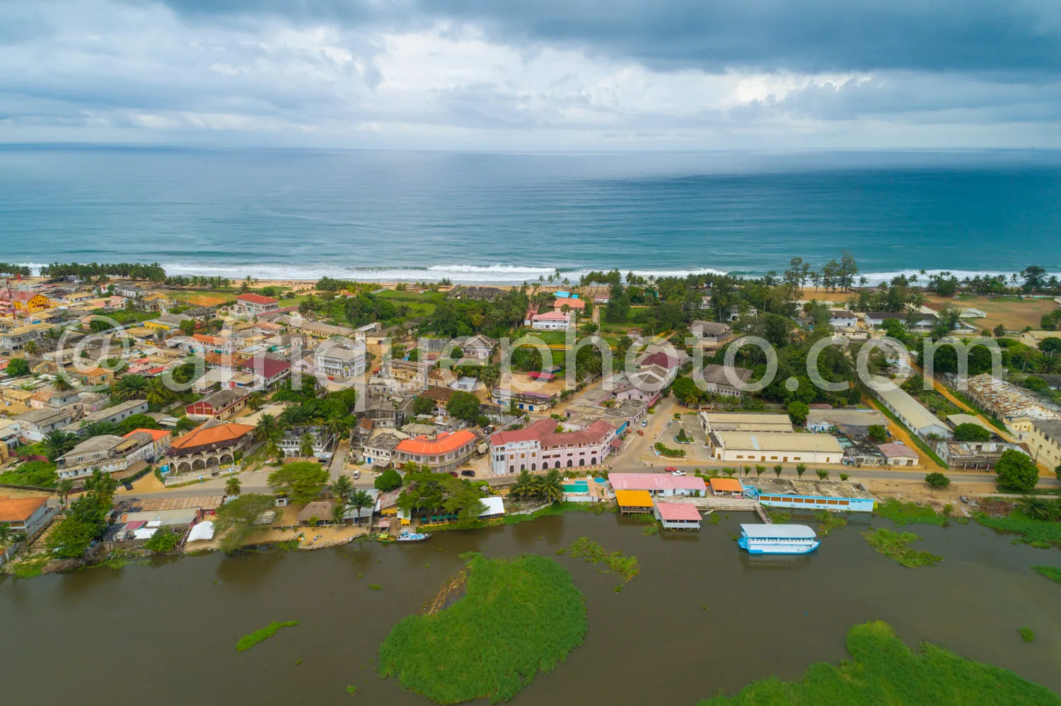 Africa photo — Vue aérienne d'un village côtier avec des maisons aux toits colorés, un embarcadère en bordure d'un plan d'eau calme et, à l'horizon, u