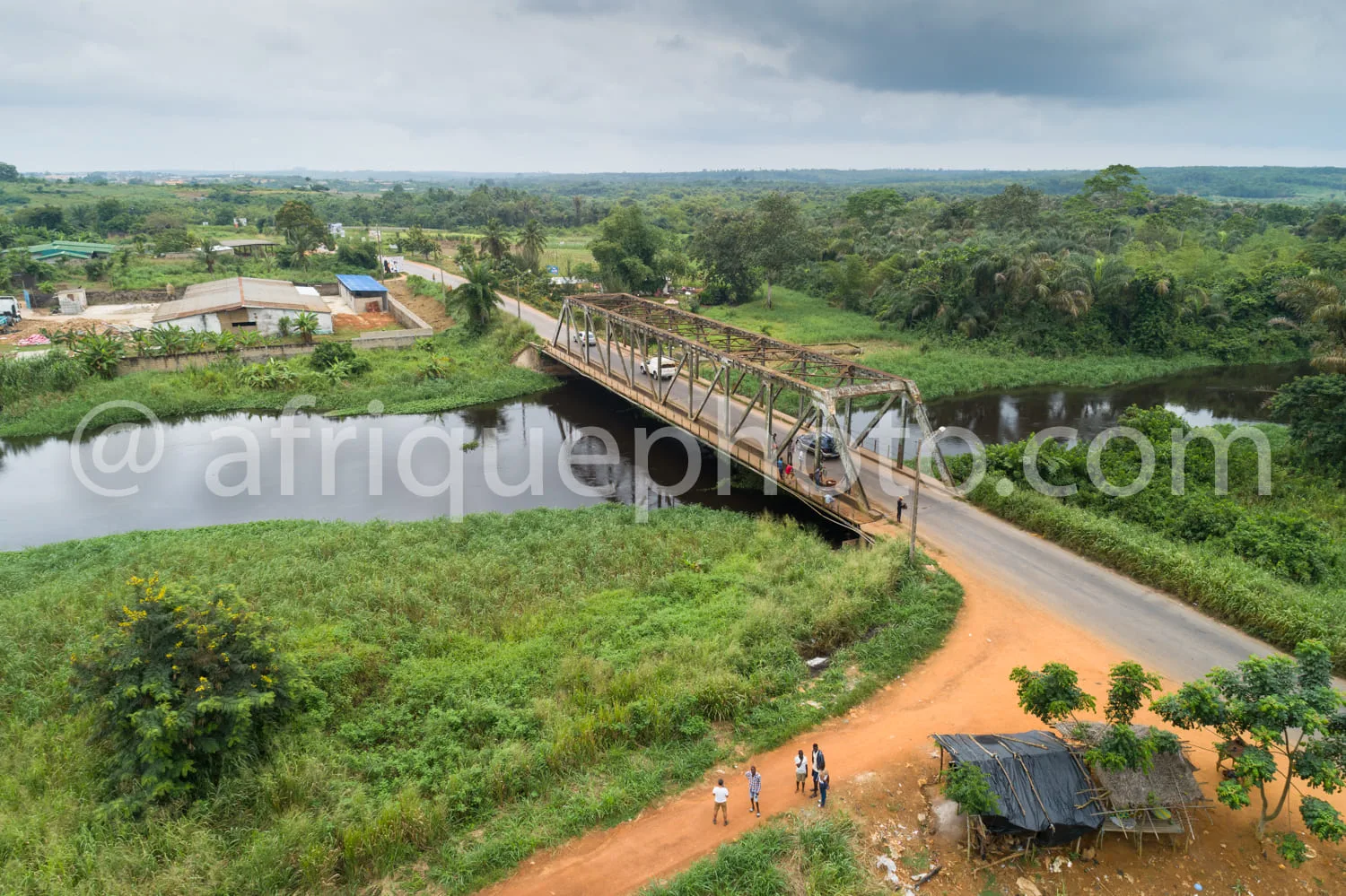 Africa photo — Vue aérienne d’un pont métallique enjambant une rivière calme, entouré de végétation luxuriante. À gauche, des maisons en dur et des ch