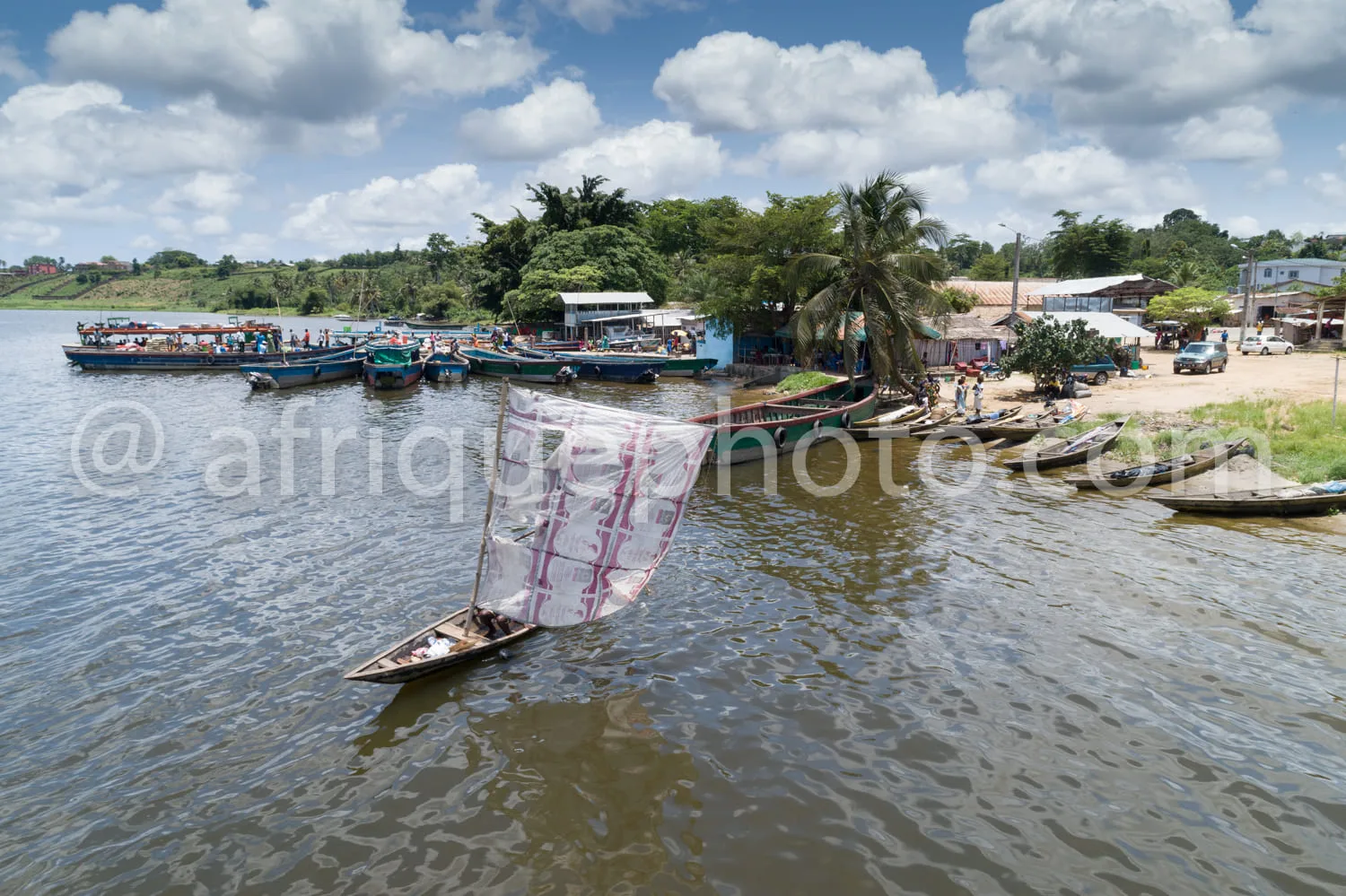 Africa photo — Vue aérienne d’un port fluvial actif où des pirogues et bateaux sont amarrés le long d’une rive ombragée par des palmiers et des arbres