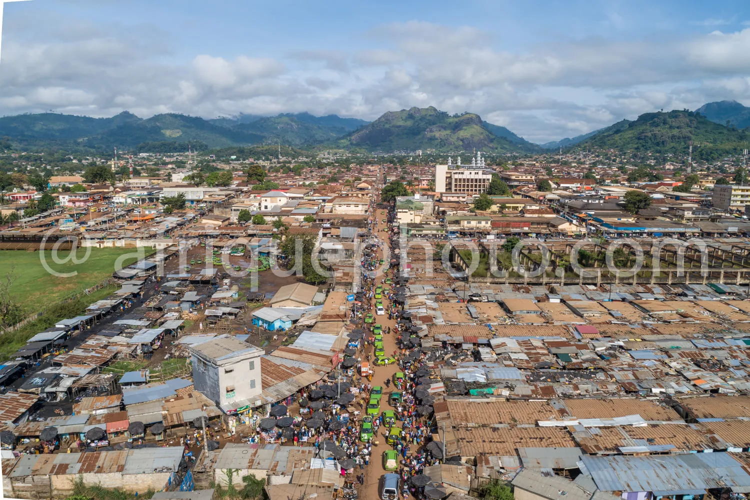Africa photo — Image montrant une vue aérienne d’un grand marché traditionnel très fréquenté, avec des étals sous abris en tôle ondulée, une rue centr
