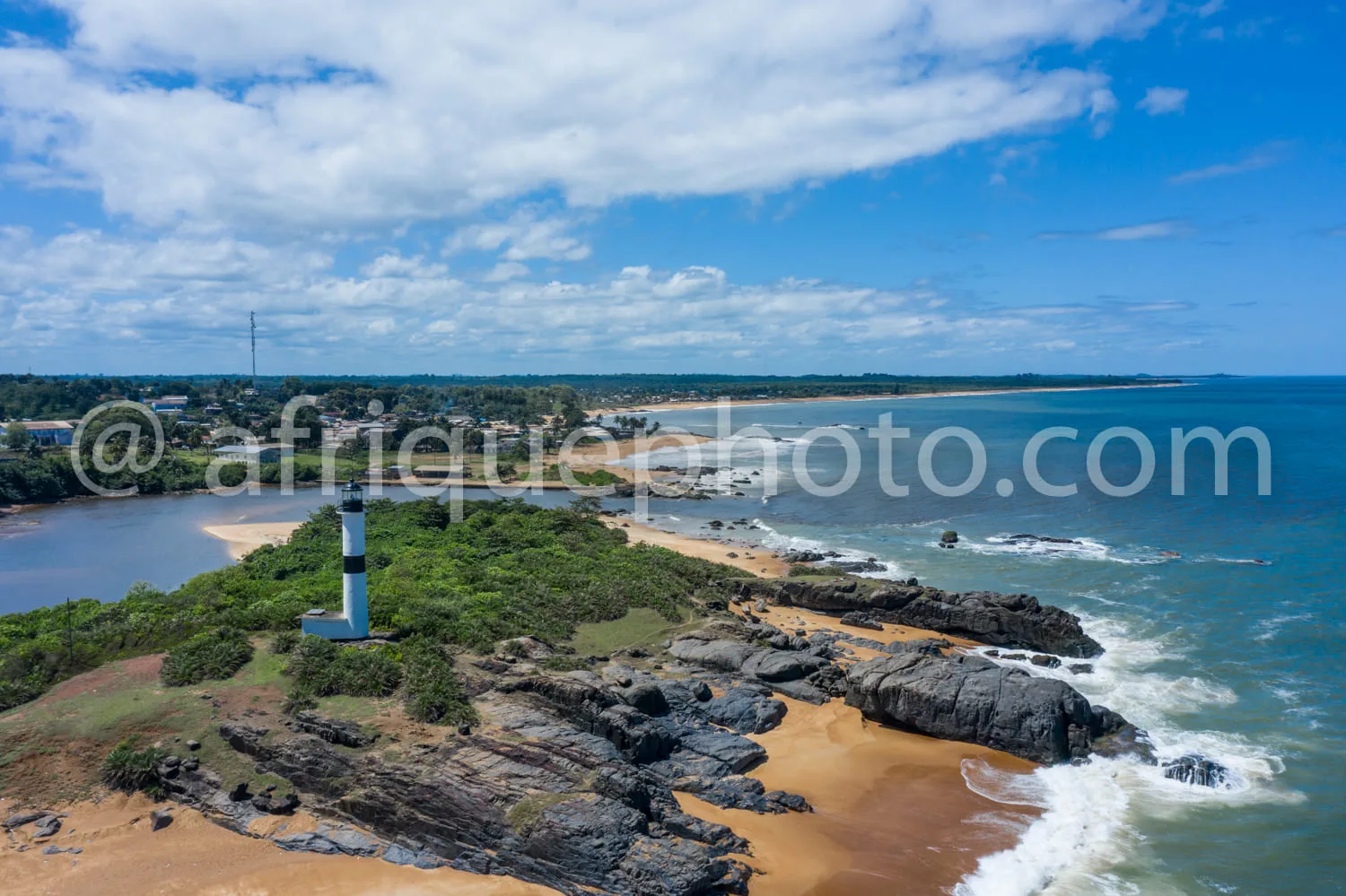 Africa photo — Un phare blanc et noir se dresse sur une petite pointe rocheuse entourée de végétation, face à une plage de sable doré bordée d’eaux tu