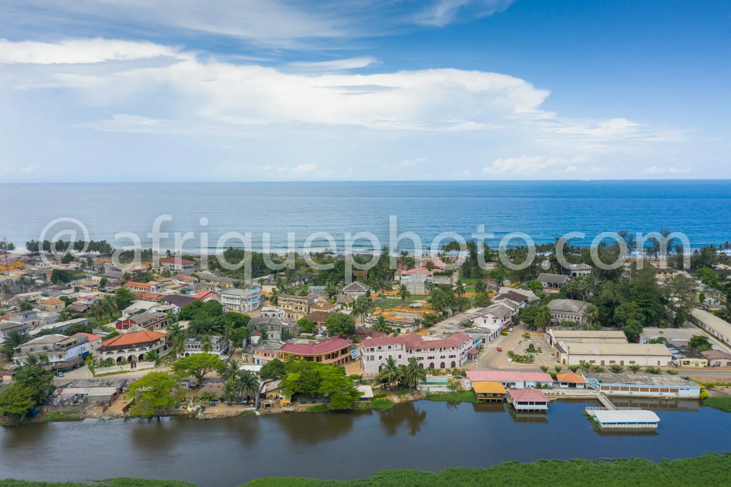 Africa photo — Vue aérienne d’un village côtier avec des maisons aux toits colorés, entourées de palmiers et de végétation dense, une lagune calme en