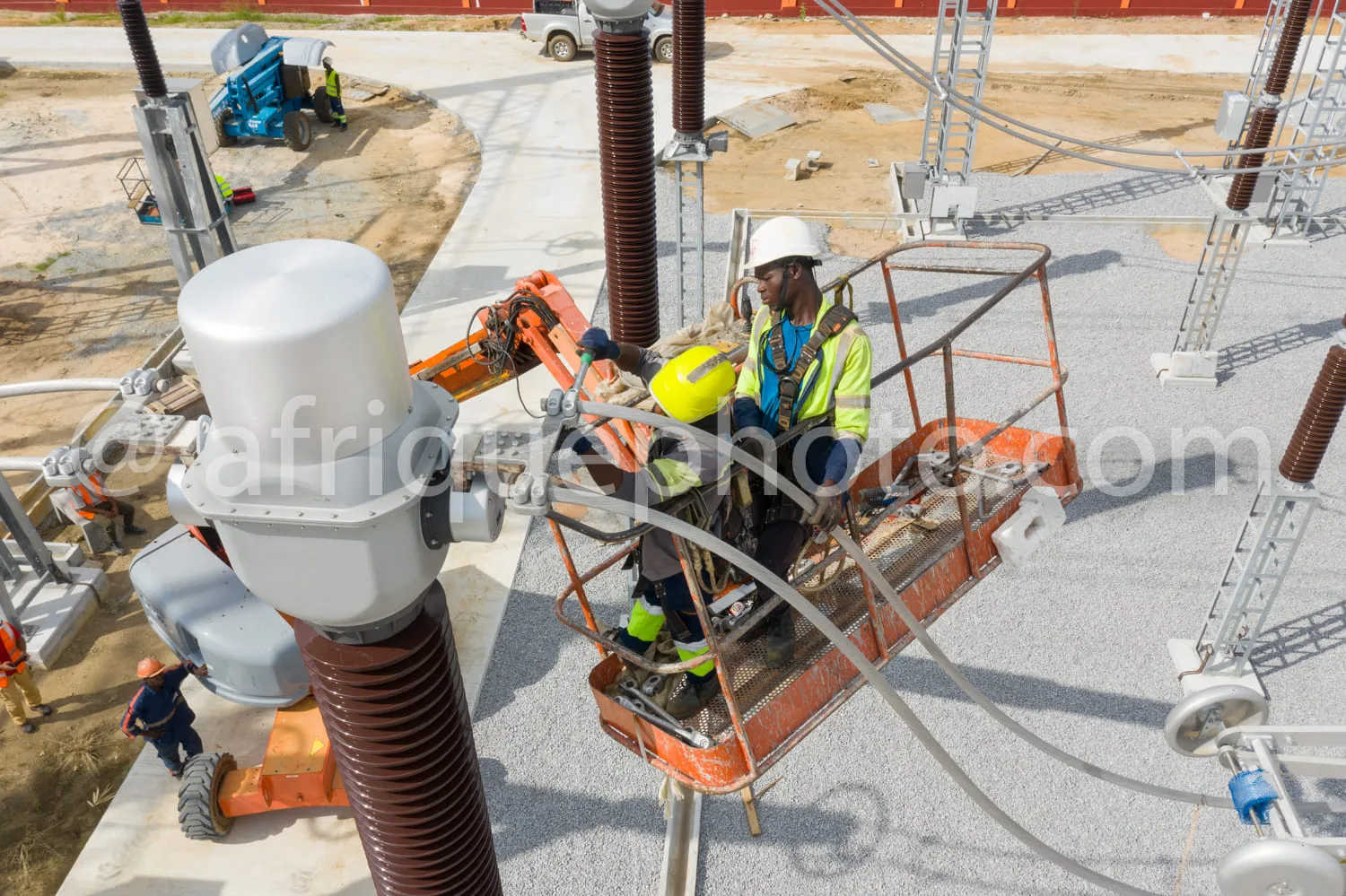 Africa photo — Un technicien en combinaison de travail jaune et casque de sécurité est assis dans une nacelle élévatrice orange, manipulant des équipe