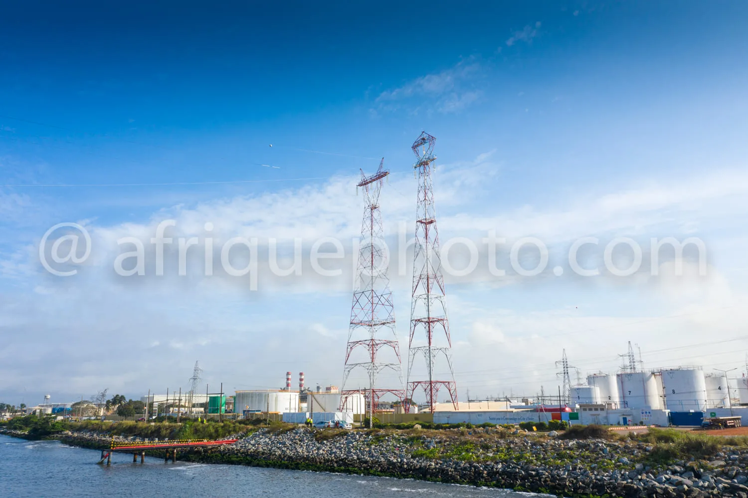 Africa photo — Vue d'une usine industrielle située près d'un plan d'eau, avec des réservoirs blancs, des bâtiments industriels et deux grands pylônes