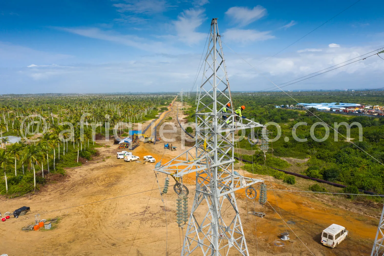 Africa photo — Vue aérienne d’un chantier de construction d’une ligne électrique haute tension traversant une zone rurale avec des palmiers, des véhic