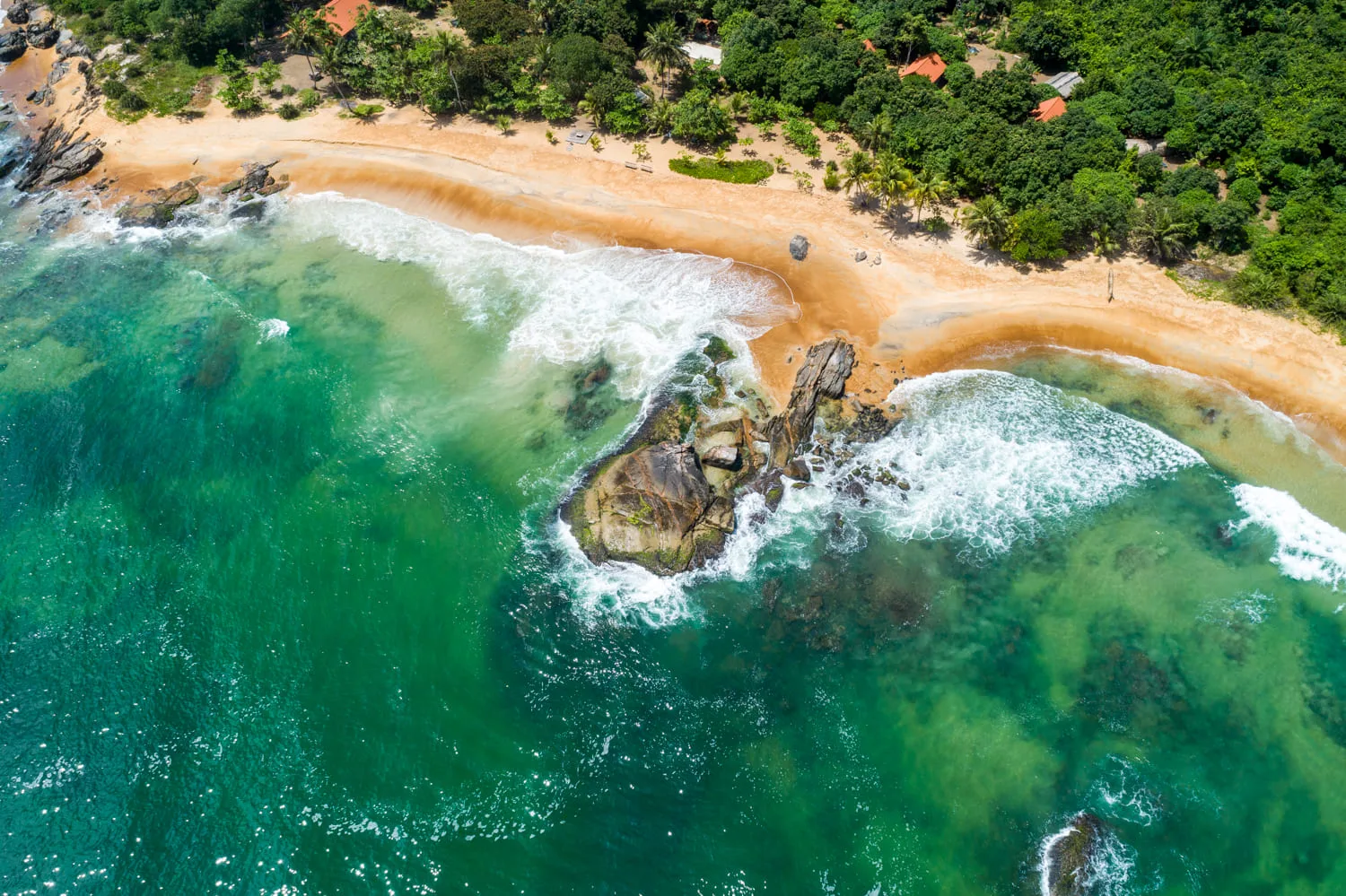 Africa photo — Vue aérienne d’une plage de sable fin bordée d’une eau turquoise aux teintes profondes, avec deux rochers émergents couverts de végétat