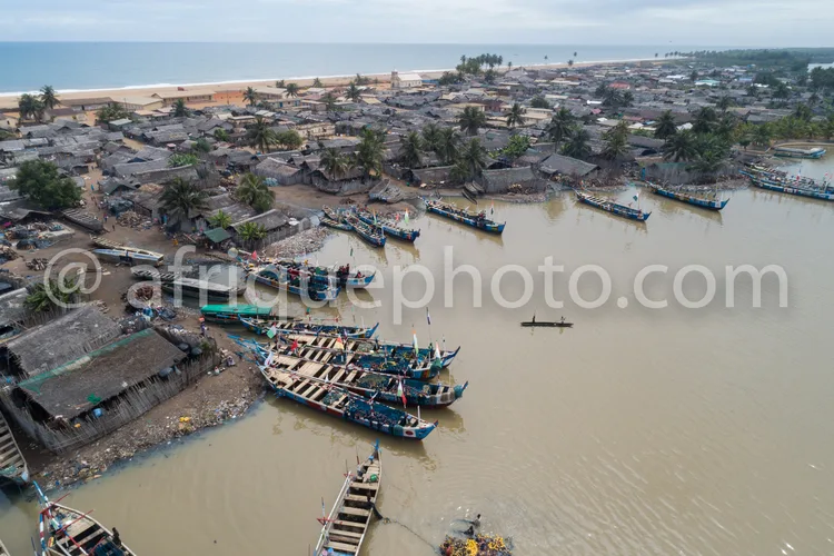 Africa photo — Vue aérienne d’un port fluvial animé où des bateaux de pêcheurs sont amarrés le long d’un fleuve. En arrière-plan, des maisons en matér