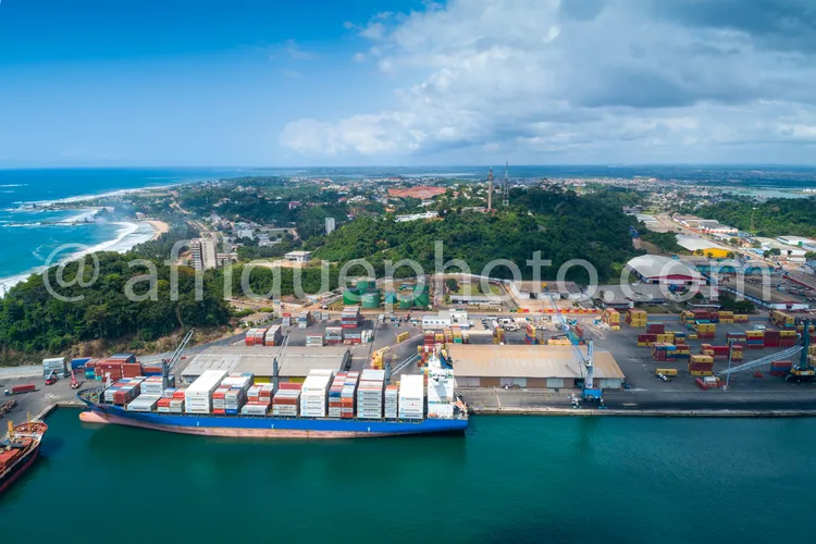 Africa photo — Vue aérienne d’un port maritime où un grand navire cargo chargé de conteneurs colorés est amarré près d’un quai. À l’arrière-plan, on a