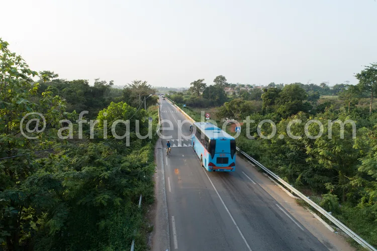 Africa photo — Un bus turquoise circule sur une route asphaltée à deux voies, bordée de végétation dense des deux côtés, sous un ciel légèrement voilé