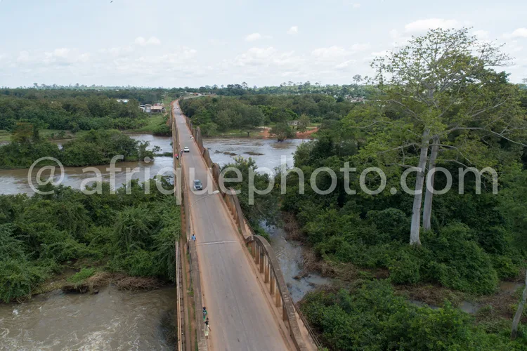 Africa photo — Vue aérienne d’un pont en béton traversant une rivière peu profonde, entouré de végétation dense et de champs. Deux véhicules et un cyc