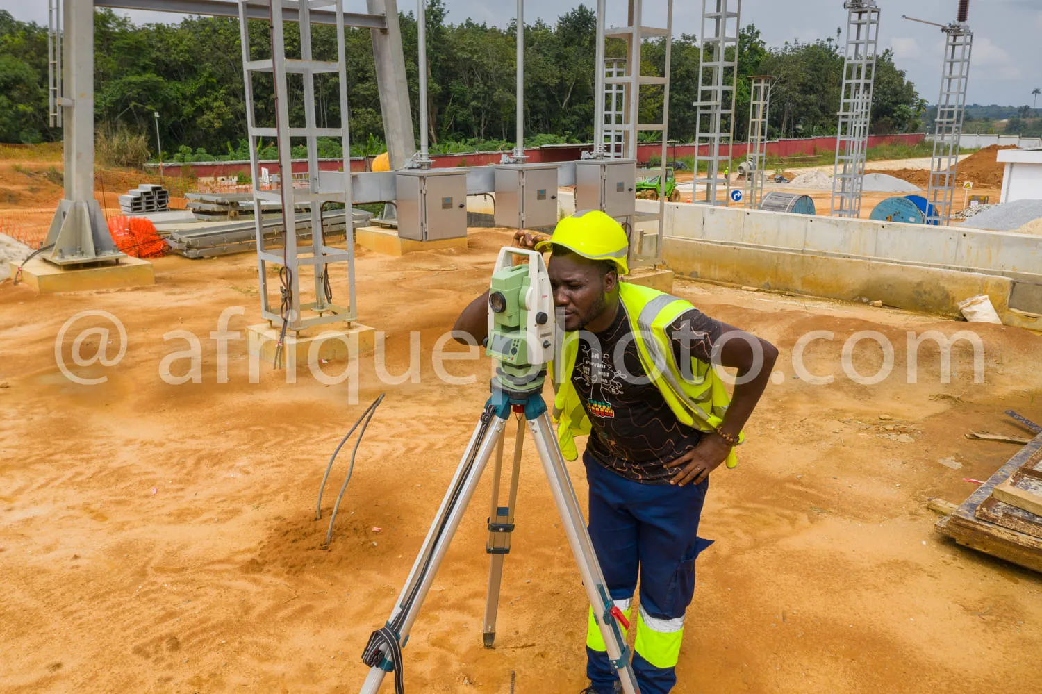 Africa photo — Un professionnel en tenue de sécurité jaune et casque, debout sur un sol terreux, manipule un théodolite posé sur un trépied pour des r