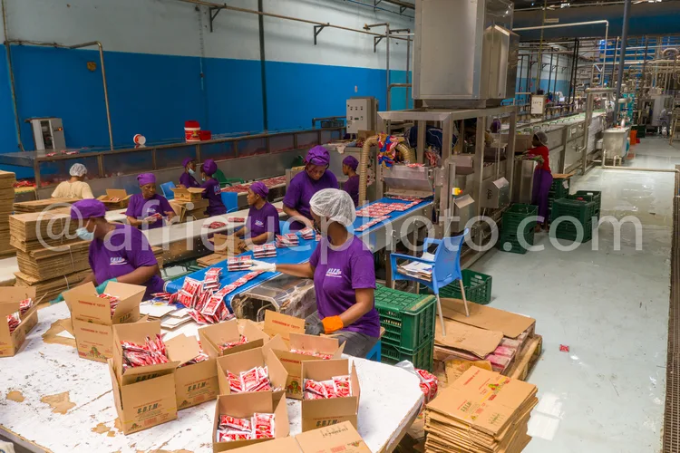 Africa photo — Une équipe de six personnes en tenue de travail, coiffes et gants violets, emballe des produits alimentaires sur une table d’assemblage