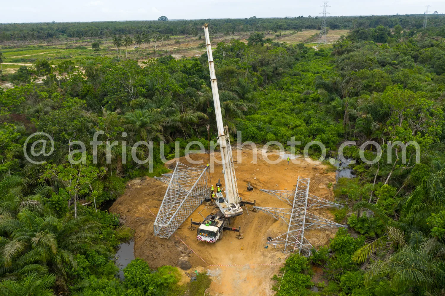 Africa photo — Vue aérienne d’une grue de construction installant une tour électrique métallique dans une zone de forêt tropicale dense, entourée de v