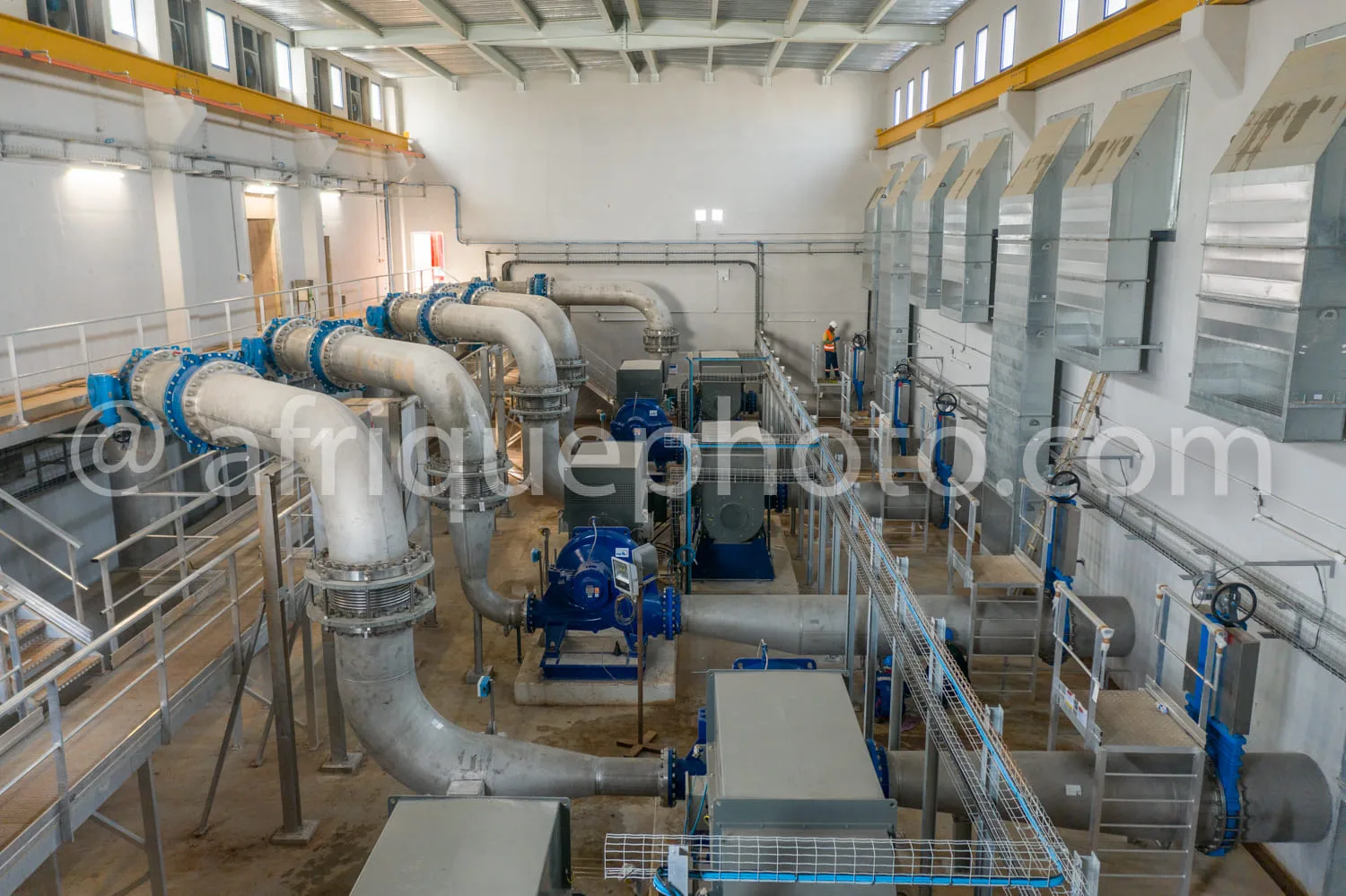 Africa photo — Vue intérieure d'une usine industrielle montrant un vaste réseau de tuyauteries en métal, des ventilateurs industriels bleus et gris, d