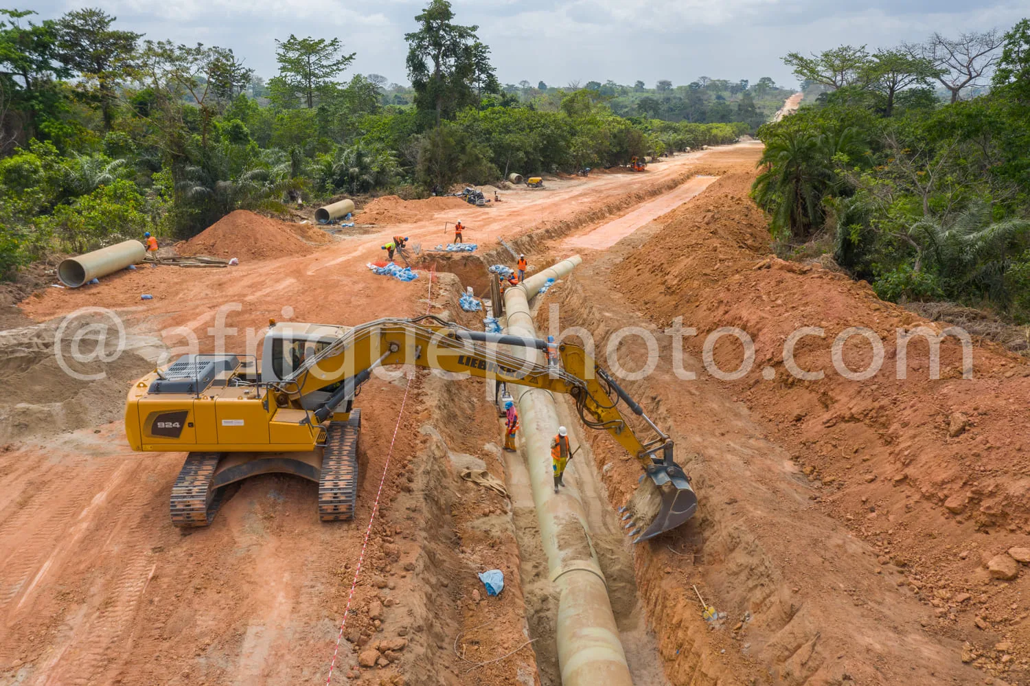 Africa photo — Image montrant un chantier de pose de canalisations en terre rouge, avec une pelle mécanique jaune positionnée pour manipuler de grands