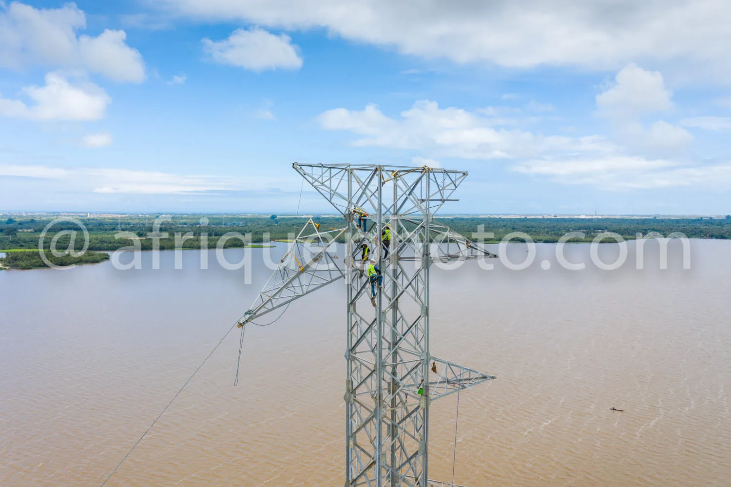 Africa photo — Deux techniciens en équipement de sécurité travaillent en hauteur sur un pylône électrique métallique situé au bord d’un large fleuve a