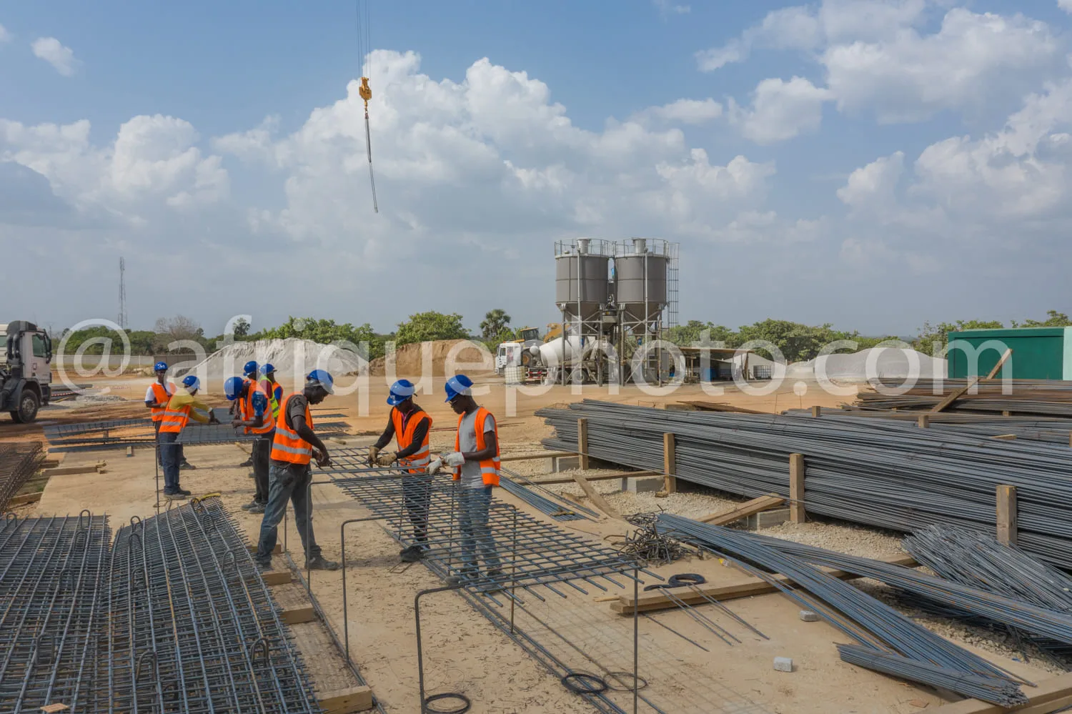 Africa photo — Cinq ouvriers en gilets orange et casques bleus travaillent sur un chantier de construction, assemblant des armatures métalliques pour
