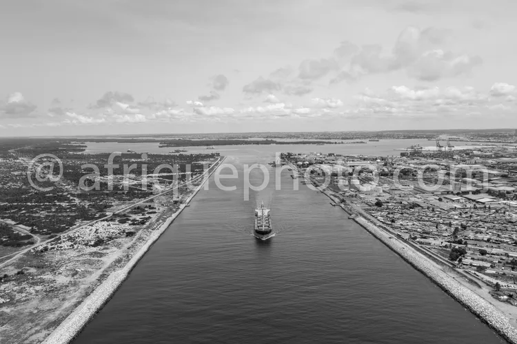 Africa photo — Un navire cargo de taille moyenne navigue au centre d’un canal bordé de digues en béton, avec des zones urbaines et industrielles de ch