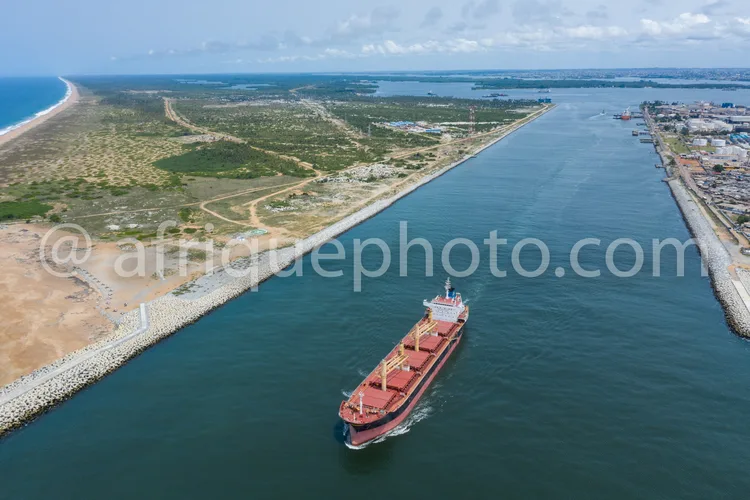 Africa photo — Un grand pétrolier rouge se déplace sur un canal maritime encadré par des digues en béton. À gauche, une plage de sable et une végétati