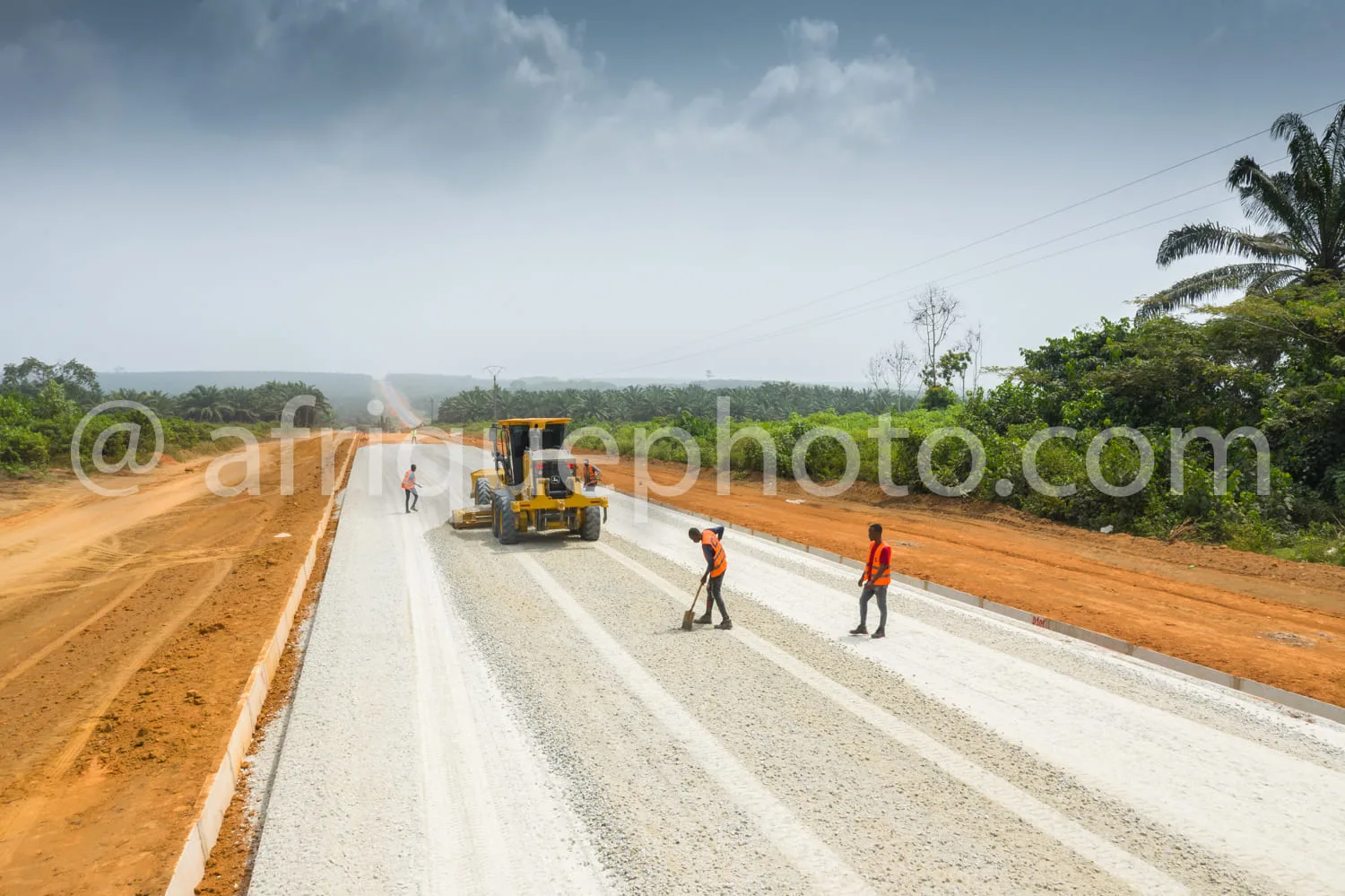 Africa photo — Une route en construction avec trois ouvriers en gilets de sécurité travaillant sur le bas-côté, un engin jaune de nivelage en action a