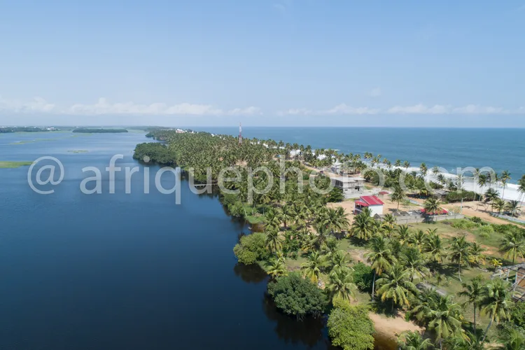 Africa photo — Vue aérienne d’un village tropical avec une longue rangée de palmiers bordant une lagune calme, des maisons colorées et un accès à la m