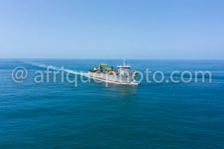 Africa photo — Un bateau de pêche de taille moyenne navigue sur une mer calme et bleue sous un ciel clair, laissant derrière lui une traînée d’eau. Le
