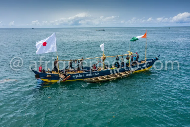 Africa photo — Un bateau coloré nommé Nyebre Sesayo navigant sur une mer calme, arborant trois drapeaux : celui du Japon à gauche, un drapeau blanc au