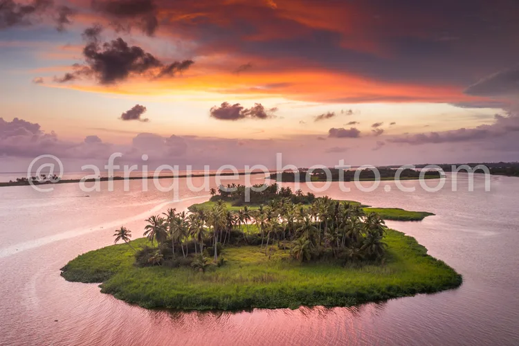 Africa photo — Une petite île entourée d’eau calme, avec une végétation luxuriante de palmiers et d’arbustes, sous un ciel aux teintes roses et orange