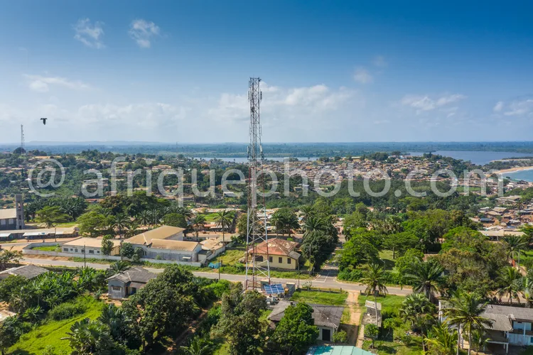Africa photo — Vue aérienne d’une ville côtière avec deux grandes antennes de télécommunication au centre, entourée de maisons, de routes sinueuses, d