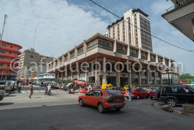 Africa photo — Un grand immeuble de style architectural années 70, à plusieurs étages, situé en plein cœur d’une rue animée. On y voit des taxis rouge