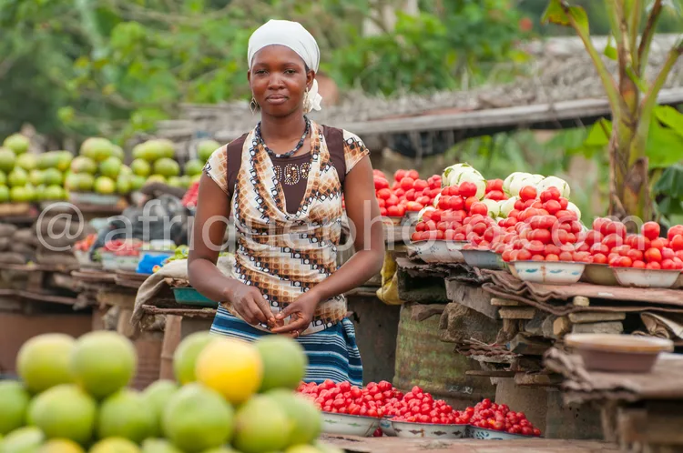 Africa photo — Une femme souriante, vêtue d’un chemisier imprimé et d’un foulard blanc sur la tête, pose devant un étalage de fruits variés. À gauche,