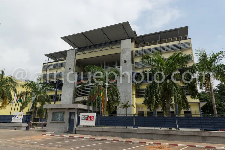 Africa photo — Un immeuble moderne de deux étages avec une façade en béton clair et des balcons en métal noir. Des plantes tropicales, dont des palmie
