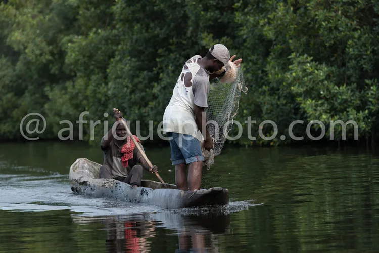 Africa photo — Deux hommes sont assis dans une pirogue en bois sur un plan d’eau calme, entourée de végétation dense. L’un d’eux tient un filet de pêc