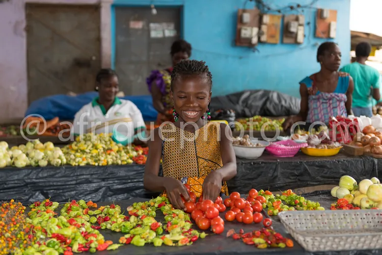 Africa photo — Une jeune fille souriante, vêtue d’une robe traditionnelle à motifs, trie des tomates rouges et des légumes variés sur une table de mar