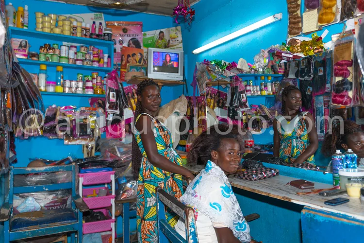 Africa photo — Intérieur d’un salon de coiffure aux murs bleus où deux femmes en tenues traditionnelles colorées s’occupent d’une cliente assise, ento