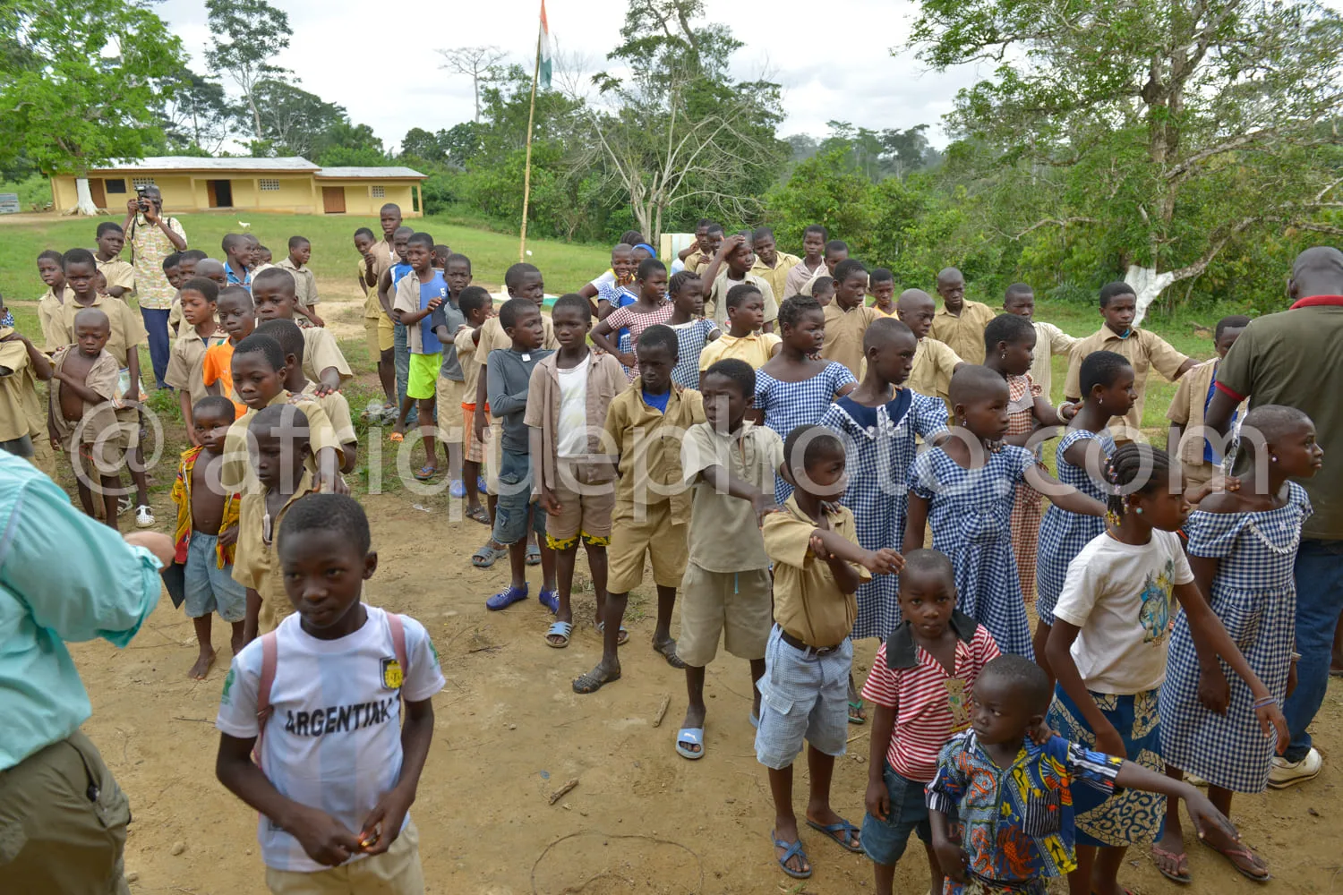 Africa photo — Un groupe d'environ cinquante enfants, majoritairement vêtus d'uniformes scolaires beiges et bleus, se tient debout sur un terrain en t