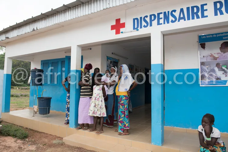 Africa photo — Groupe de femmes vêtues de robes colorées attendant devant l'entrée d'un dispensaire rural peint en bleu et blanc, avec un panneau indi