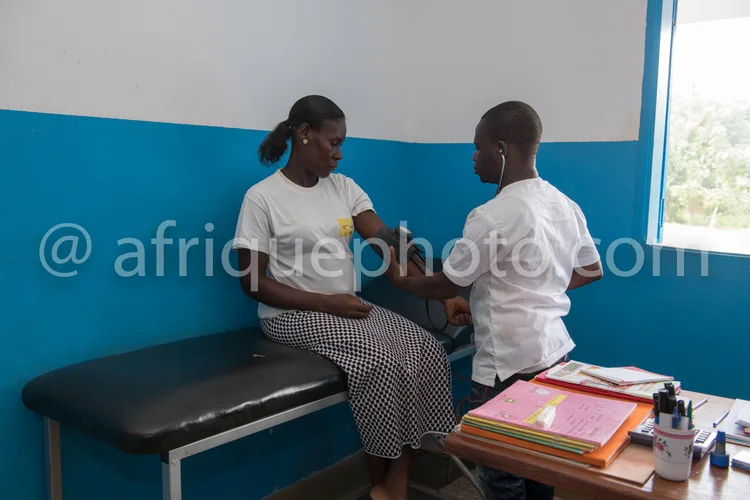 Africa photo — Une femme assise sur une table d'examen médical, un professionnel de santé en blouse blanche prend sa tension artérielle avec un tensio