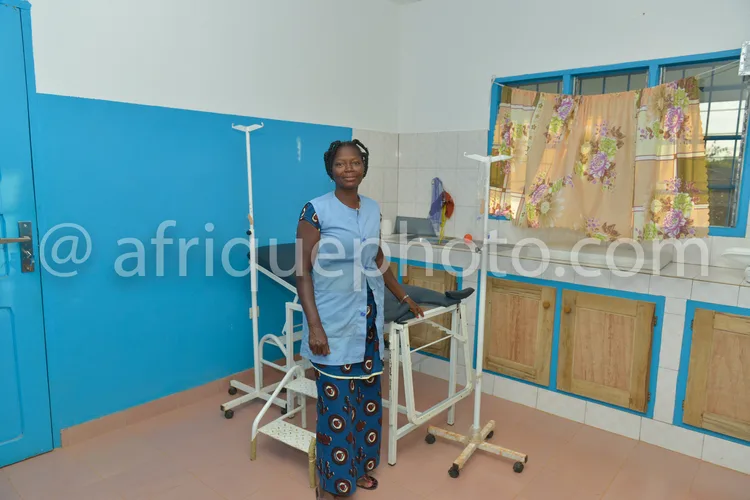 Africa photo — Une femme souriante se tient debout dans une pièce médicale, s’appuyant sur un cadre de marche blanc à quatre roues, entourée de murs b
