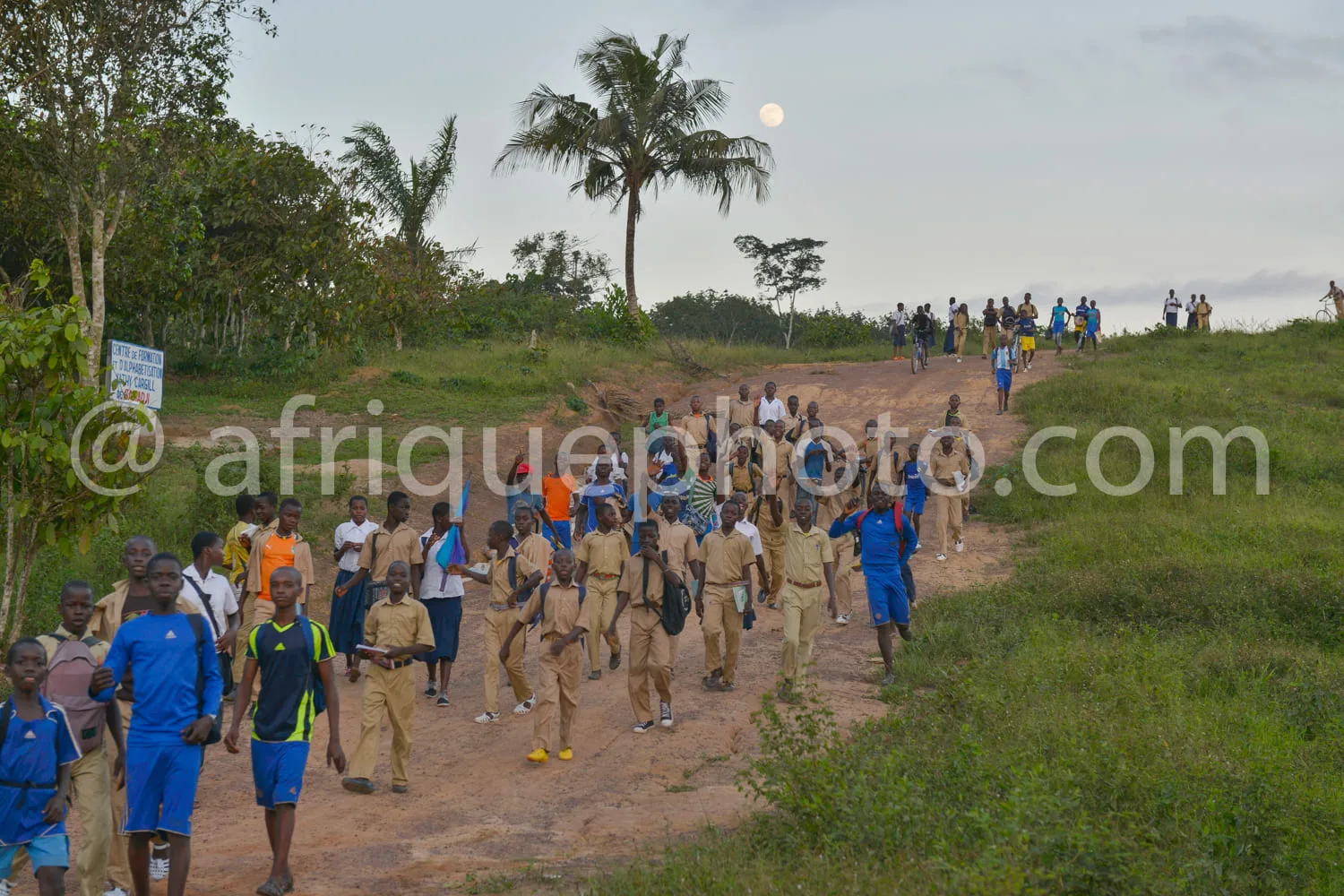 Africa photo — Un groupe d'élèves en uniforme scolaire, certains en tenue beige et d'autres en bleu, défilent sur un chemin de terre bordé de végétati