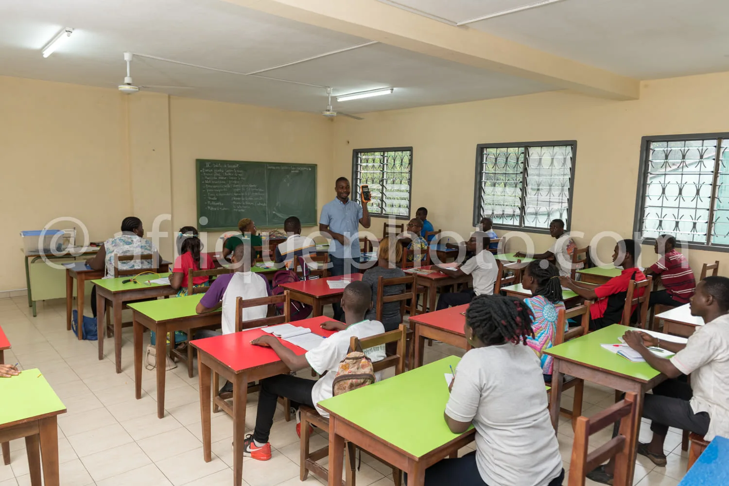 Africa photo — Une salle de classe lumineuse où un enseignant debout devant un tableau noir explique un cours à une vingtaine d'élèves assis à des bur